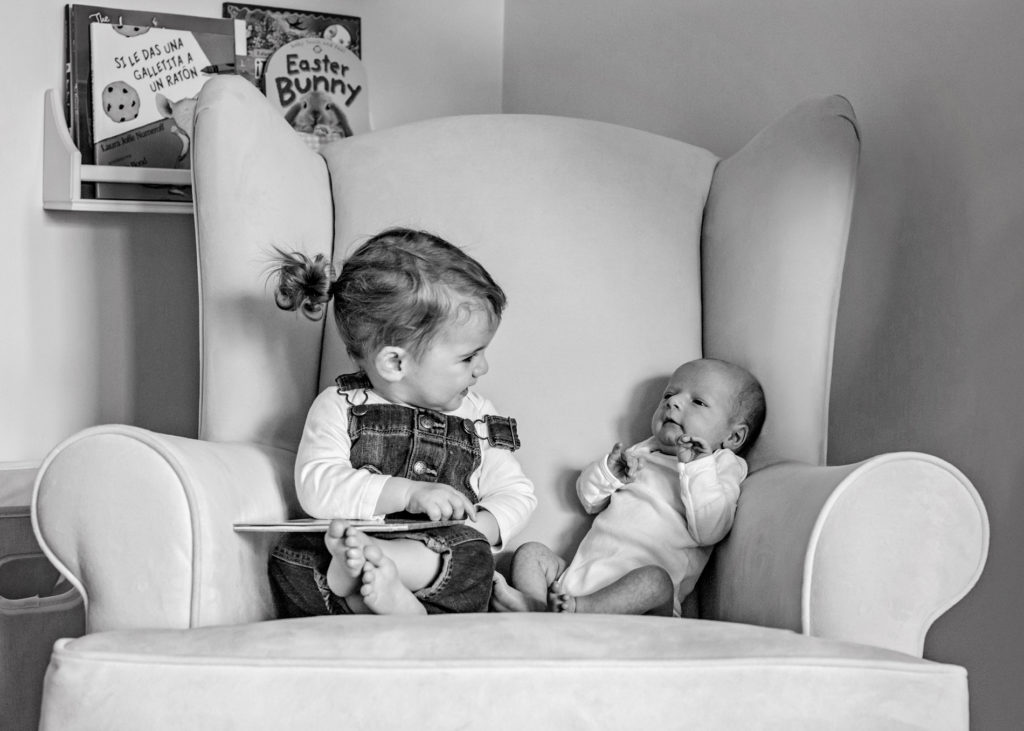 Family Photographer, older sister admires newborn sibling beside her in a chair
