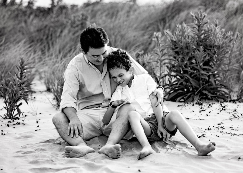 Family Photographer, son sits with dad in the beach sand