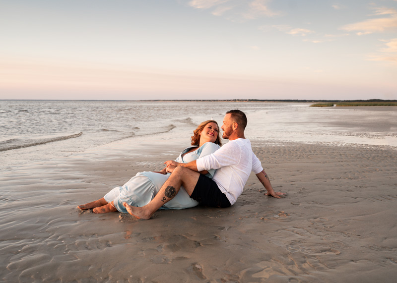 Family Photographer, man and woman admire each other while sitting in the sand at the beacg